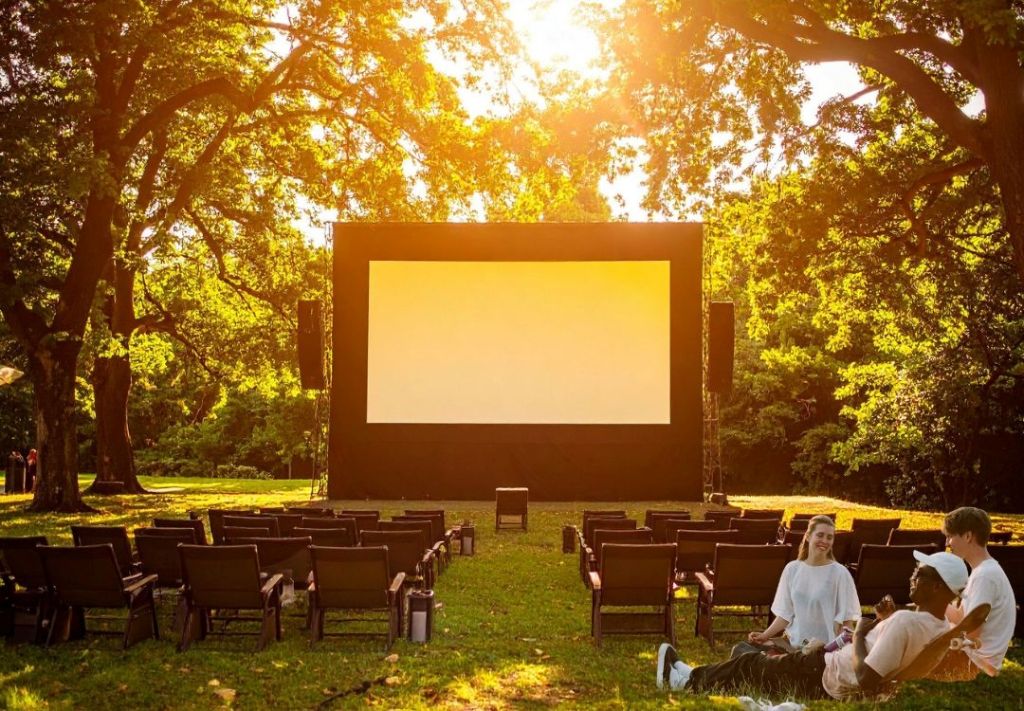 Outdoor movie theater in a lush, sunlit forest with rows of empty chairs facing a large projection screen.