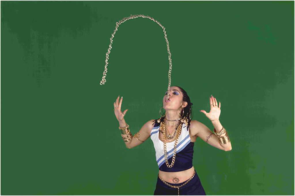 A person wearing jewelry and a crop top dancing with their hands raised in the air against a green background.