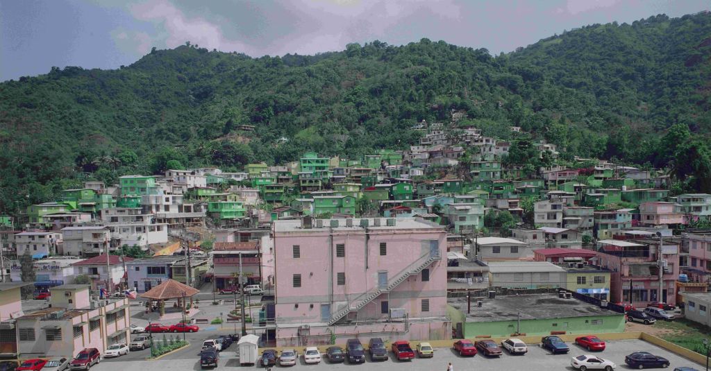 Densely packed hillside town with colorful buildings and vehicles, surrounded by lush green mountains.