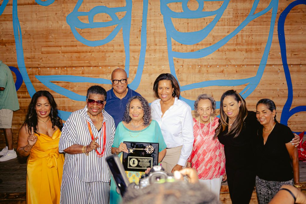 A group of diverse people posing together, some holding an award or plaque, in front of a colorful mural wall.
