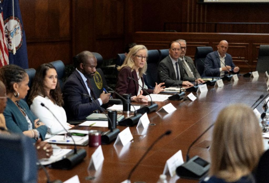 A group of people seated around a conference table in a government or legal setting, with the American flag visible in the background.
