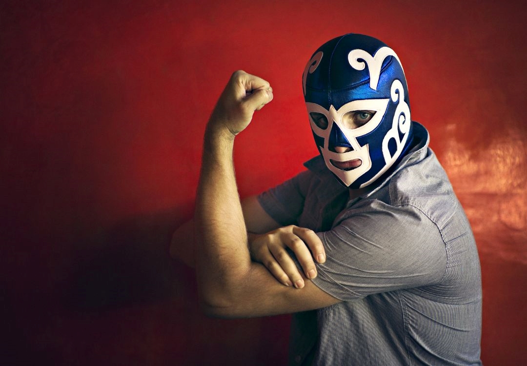 A person wearing a blue and white Mexican wrestling mask flexing their muscular arm against a red background.