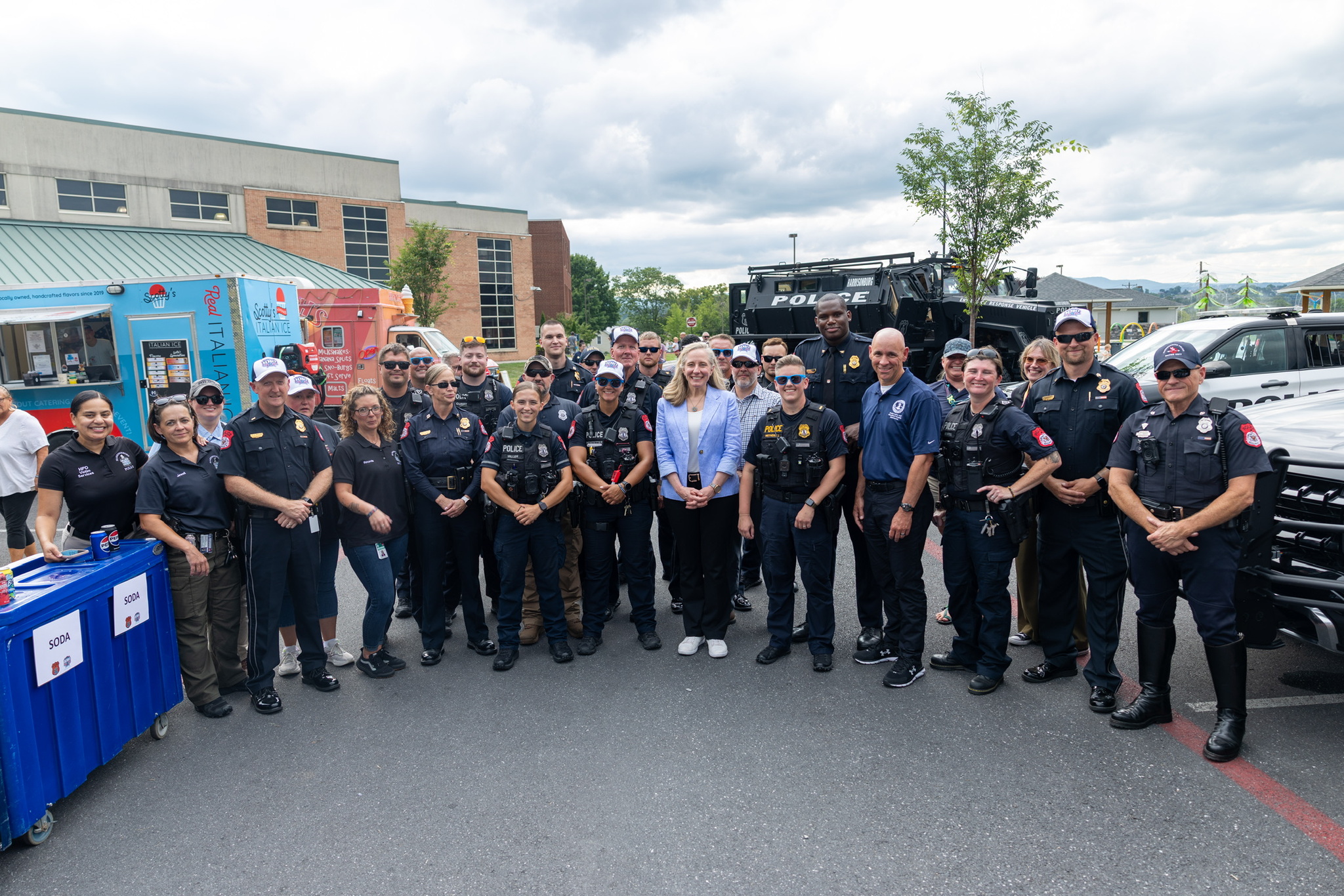 A group of police officers and staff posing for a photo outdoors, with a police vehicle and tents visible in the background.