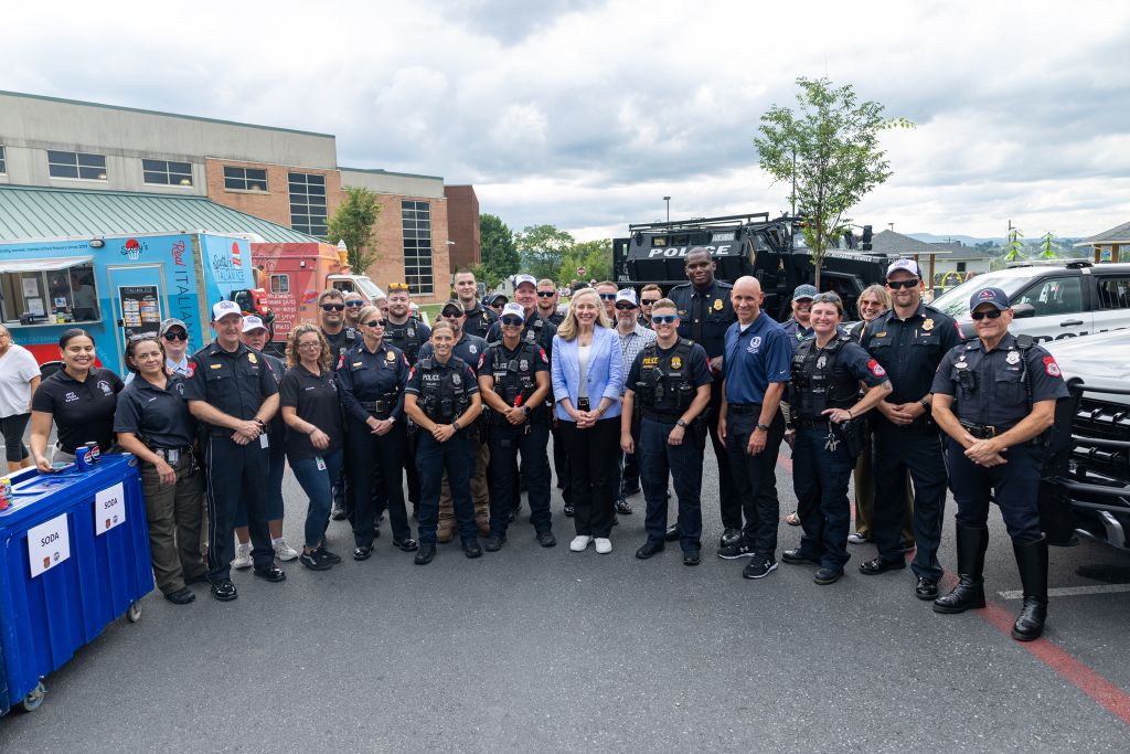A group of police officers and staff posing for a photo outdoors, with a police vehicle and tents visible in the background.