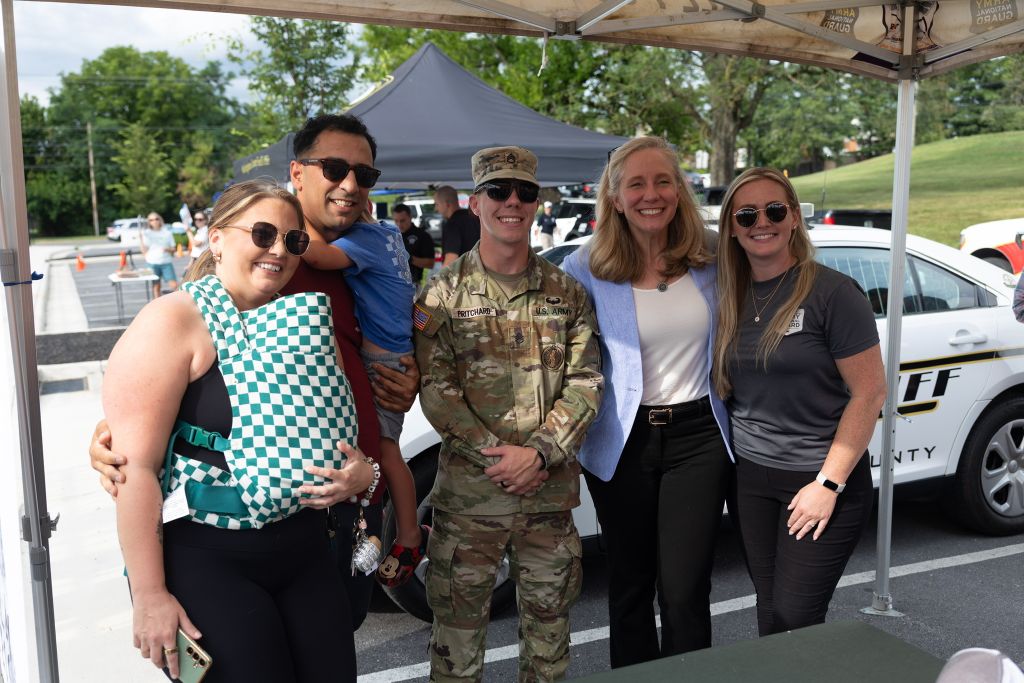 A group of five people, including a person in military uniform, posing together outdoors in front of a tent and police car.