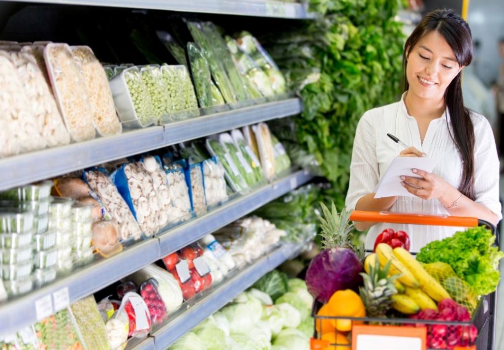 A smiling woman holding a clipboard while shopping in a grocery store aisle filled with fresh produce and packaged goods.